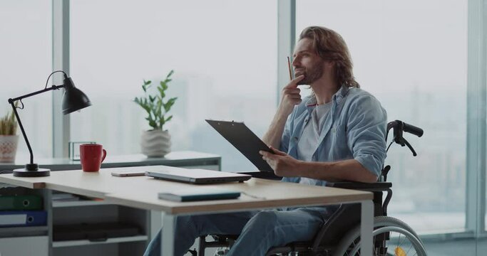 Disabled young male office executive sitting in wheelchair at desk. Corporate person with disabilities writing notes performing work duties in the office.