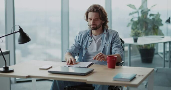 Disabled invalid young male designer working in corporate office, developing creative ideas and drinking coffee. Freelance lifestyle. Corporate person.