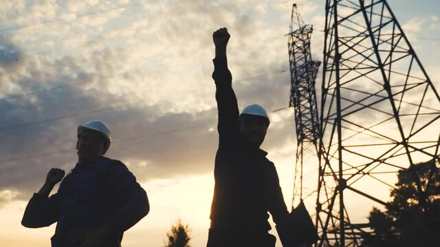 silhouette of an electricians engineers success next to an electrical support tower energy. electricity teamwork tower support. electricals engineers success sunset teamwork electricity pylon