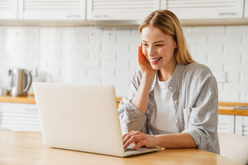 Joyful blonde girl smiling and using laptop while sitting at kitchen