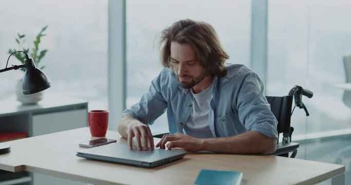 Frustrated young hesitating man using computer laptop sitting by desk doubting what to do. Self-doubting disabled man in wheelchair working first day at office.
