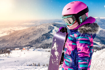 Fototapeta na wymiar Little girl snowboarding with equipment helmet and goggles outwear holding snowboard resting on top of ski slope in sunlight