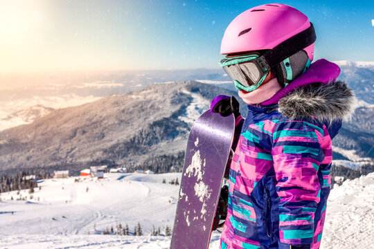 Little girl snowboarding with equipment helmet and goggles outwear holding snowboard resting on top of ski slope in sunlight