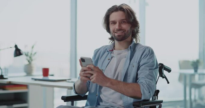 Optimistic young caucasian office worker male using mobile phone social media smiling to camera inside corporate coworking workspace. Portraits.