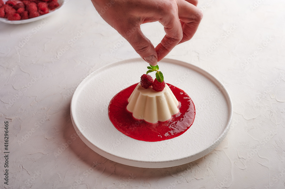 Wall mural Woman's hand puts mint leaf to panna cotta with syrup and berries, high key, close up