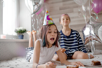 Two preteen sisters twins celebrate birthday day at the morning, active kids dressed in pajamas have fun in the bed with balloons. Children eat cake in the bed