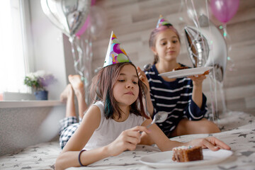 Two preteen sisters twins celebrate birthday day at the morning, active kids dressed in pajamas have fun in the bed with balloons. Children eat cake in the bed