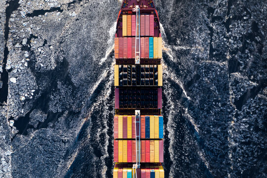 Cargo ship driving through frozen sea with colorful containers. Dark waters with ice blocks floating in sea. 