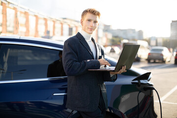 Portrait of a stylish young Caucasian businessman standing near charging electric car, using a laptop computer outdoors in the city EV charging station