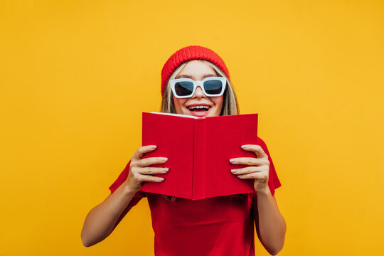 Modern cute student girl dressed in red on a yellow background, she is covered with a red book and funny looks at the camera, wearing sunglasses. Studio photo