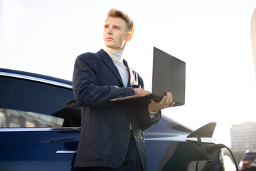 Young handsome European businessman at charging station, standing near electric car with laptop pc and looking away, while refueling his auto. EV station and people concept