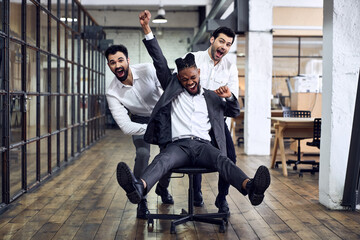 Work hard play hard. Four young cheerful business people in formal wear having fun while racing on office chairs and smiling. - obrazy, fototapety, plakaty