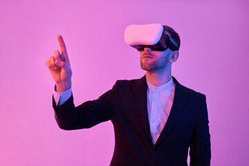 Young attractive businessman in black suit and white shirt uses virtual reality glasses, trying to press the virtual button with his finger isolated on neon pink background. Future technologies.