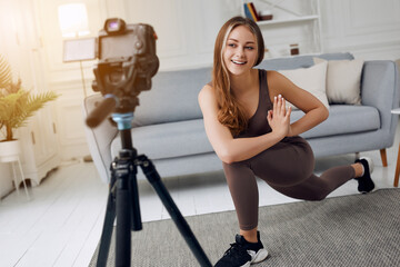 Fototapeta na wymiar Athletic woman blogger in sportswear shoots video on camera as she does exercises at home in the living room. Sport and recreation concept. Healthy lifestyle.