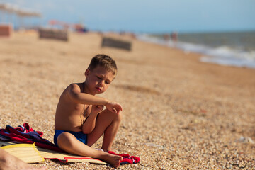 a boy sits on a red towel of a sandy beach near the sea and looks at his hand  © Татьяна Толстая
