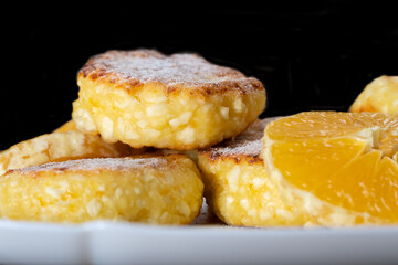 Sweet cottage cheese pancakes with orange slices on a white plate close-up against a black wall with copy space.