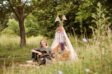 Authentic woman is playing the guitar next to wigwam outdoors in summer.