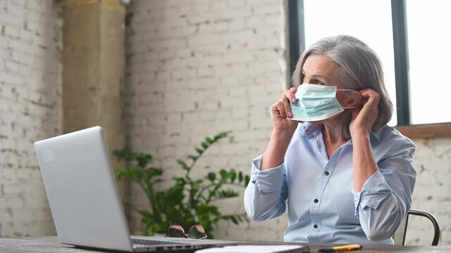 New rules for virus and flu protection for office employees. Senior mature business woman covered her face and using a laptop indoor, elderly lady staying safe with a protective face mask
