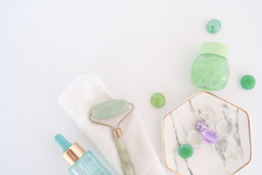 Green cream, green face roller, white towel a blue serum pot, green and violet stones for skincare
