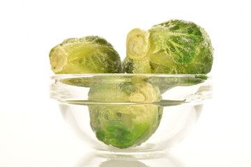 Several frozen natural Brussels sprouts in a glass bowl, close-up, isolated on white.
