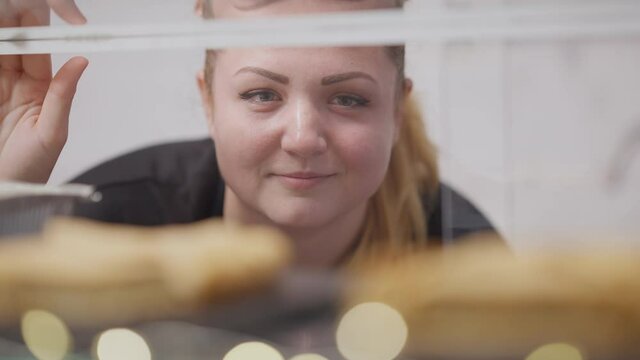 Close-up face of smiling plus-size woman looking at sweet delicious desserts lying on glass counter in cafe. Portrait of positive confident Caucasian lady choosing snack in cafeteria.