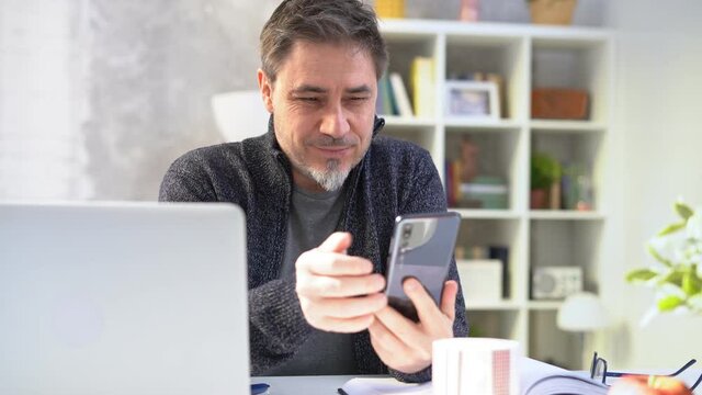 Older white man in glasses sitting at desk using phone. Home office, businessman working from home, morning coffee, checking social media news feed.