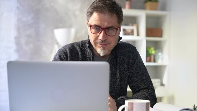 Bearded man working online with laptop computer at home sitting at desk. Home office, browsing internet, study room. Portrait of mature age, middle age, mid adult man in 50s.