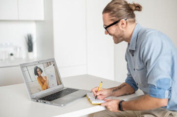 Young diligent employee with glasses and hair pulled up, sitting at the table, at a working meeting with a colleague that shows graphs, looking at the laptop screen, working online in the kitchen