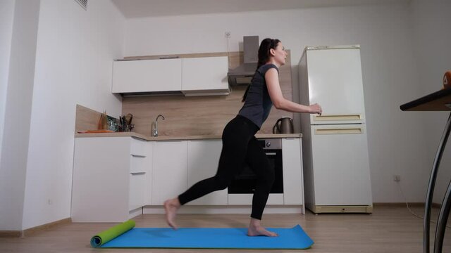 A young energetic girl in black sports leggings and a grey T-shirt will perform a squat exercise with her feet falling back standing on a blue mat against a white kitchen. High quality FullHD footage