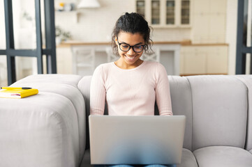 Positive intelligent African American teenage girl student in glasses sitting on the couch with laptop on the lap, studying from home, typing email, writing article, working on project, watching video