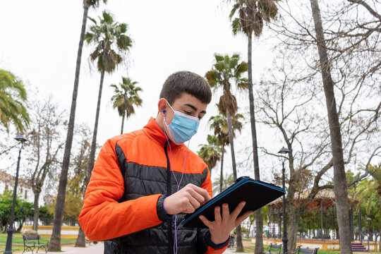 Stock photo of young boy wearing face mask due to covid19 using his tablet and listening to music in the park.