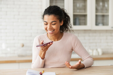 Young mixed-race ethnic teenage girl with afro hairstyle wearing casual clothes, sitting at the kitchen table and holding mobile phone, talking, asking direction, recording voice message and smiling