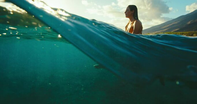Beautiful female surfer sitting on surfboard in ocean at sunset waiting for the next wave. Slow motion shot on RED cinema camera.