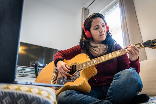Stock photo of concentrated woman playing her guitar and singing while following tutorial in her laptop.