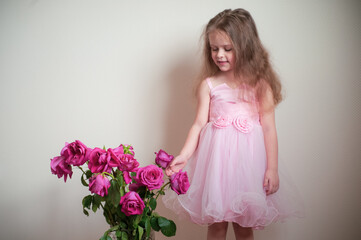 A cute little girl with long hair in a beautiful pink dress near a vase with roses that have faded a little