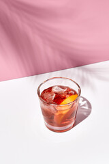Negroni cocktail over pink background. Drink in rox glass in daylight with palm leaf hard shadow. Summer, tropical, fresh drink concept