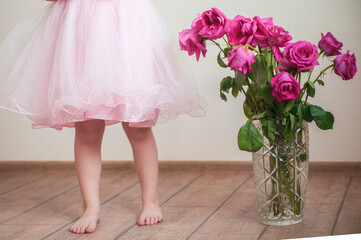 Children's feet under a lush pink dress stand near a vase with withered red roses