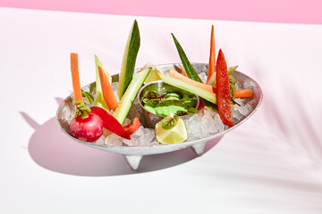 Veggie plate with crushed ice and green dip. Restaurant appetizer plate on white table with pink wall. Day sunlight with hard shadow of fern palm leaves. Summer or spring restaurant food concept.