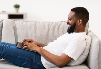 Black Freelancer Guy Working On Laptop Lying On Sofa Indoor