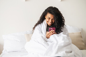 Happy young mixed-race woman girl with curly hair covered in a cozy soft blanket staying at home in...