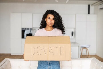 Young pretty African American woman standing in the living room and holding carton box for donation and donate is written by hand, mixed-race girl looking at camera, charity concept, sharing is caring