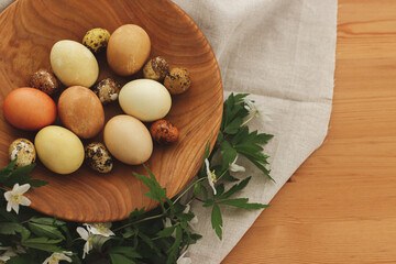 Modern easter eggs in wooden bowl with spring flowers on rustic table, top view. Happy Easter!