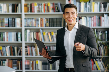 Joyful young adult hispanic freelancer or student stands in modern office or library, using laptop. Happy male employee in stylish formal suit posing, looks at the camera, smiling