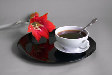 On a gray background, a composition of a cup of coffee on a black plate and a red lily