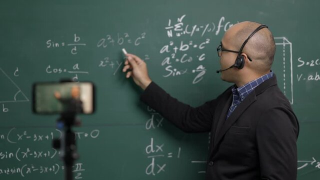 Indian young teacher man wearing headset teaching online video conference live stream by smartphone. Asian teacher teaching mathematics class webinar online for students learning.
