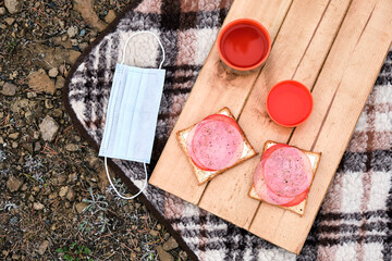 Sandwiches on the wooden stand with orange mugs from thermos with mask on the plaid. Cozy picnic on fresh air.