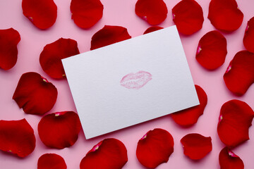 Different red petals of roses and postcard with mark of lips on pink background