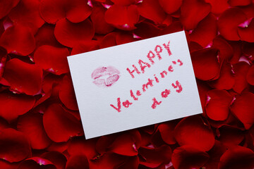 Different red petals of roses and postcard with mark of lips and lettering Happy valentine's day. Flat lay