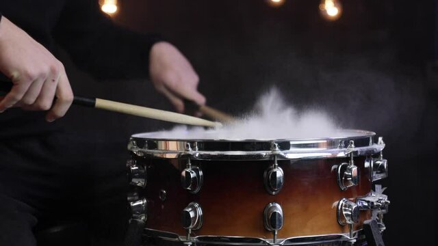 A man plays with sticks on a snare drum with smoke in dark clothes in a dark room.