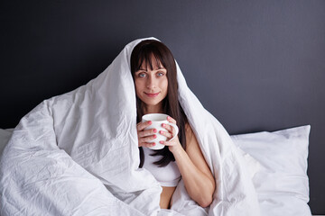 Time for myself. Comfort and relaxation. Pretty young woman drinking tea while sitting on her bed at home covering by blanket.